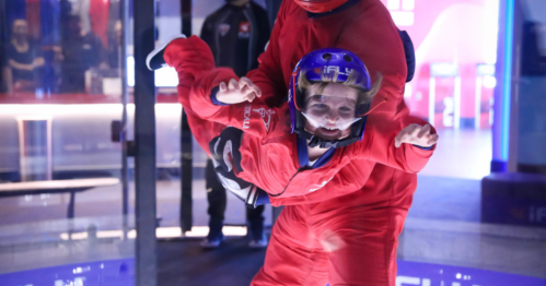 A person in a red jumpsuit enjoys indoor skydiving, being supported by an instructor in a wind tunnel.