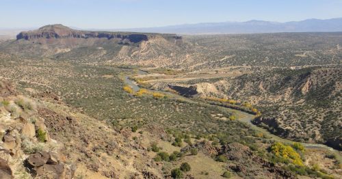A panoramic view of a rugged landscape with a winding river, rocky hills, and distant mountains under a clear sky.