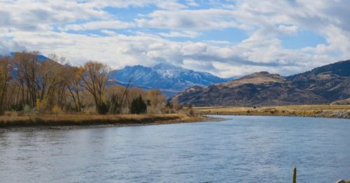 A serene river flows through a landscape of trees and mountains under a partly cloudy sky.