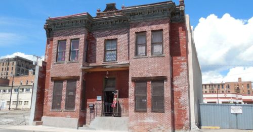 A two-story brick building with a red facade, featuring multiple windows and a small porch. Blue sky in the background.