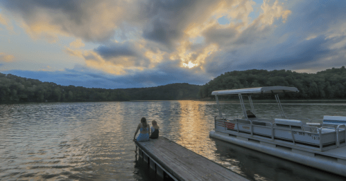 Two people sit on a dock by a lake at sunset, with a boat nearby and clouds reflecting on the water.