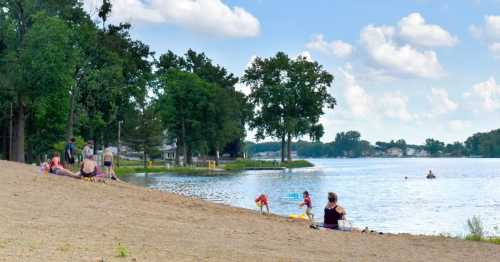 A sandy beach with people relaxing, children playing near the water, and trees lining the shore under a blue sky.