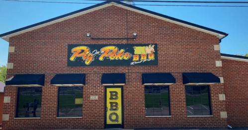 Exterior of a brick building with a sign reading "Pig 'N' Poke BBQ" and large windows, featuring a yellow "BBQ" door.