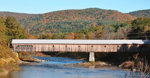 A wooden covered bridge spans a river, surrounded by colorful autumn foliage and rolling hills in the background.