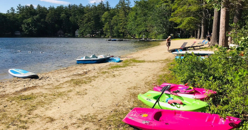 Colorful kayaks line a sandy beach by a calm lake, with trees and boats in the background under a clear blue sky.