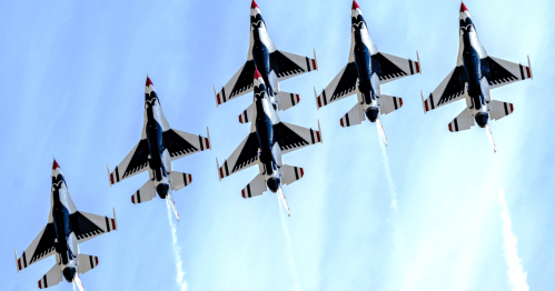 Six fighter jets flying in formation against a clear blue sky, leaving trails of smoke behind them.