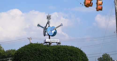 A blue guitar sign atop a bush, marking the intersection of Highway 61, with traffic lights in the background.