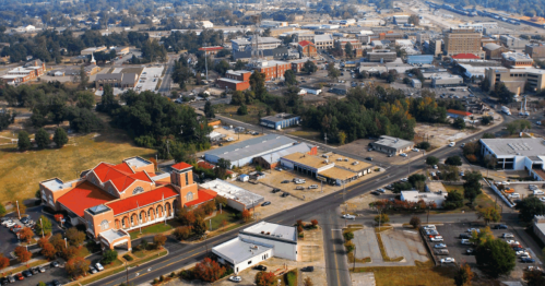 Aerial view of a cityscape featuring buildings, roads, and greenery, with a prominent red-roofed structure in the foreground.