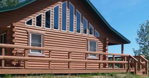 A log cabin with large windows and a green roof, surrounded by trees and blue sky.