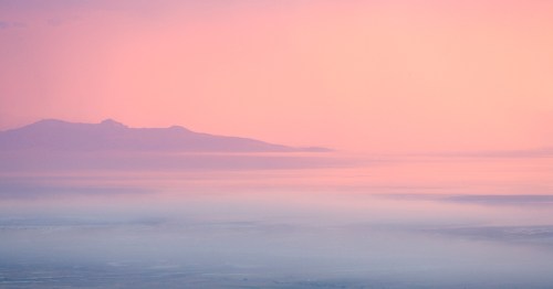 A serene landscape featuring soft pink hues over distant mountains and a calm sea, creating a tranquil atmosphere.
