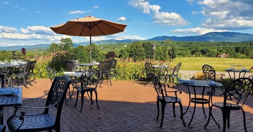 Outdoor dining area with metal tables and chairs, an umbrella, and a scenic view of mountains and blue sky.
