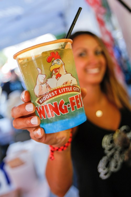 A woman holds a colorful drink in a cup featuring a chicken logo, smiling at a festival setting.