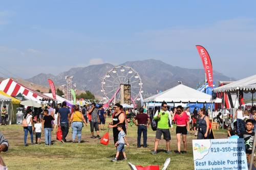 A lively outdoor fair with colorful tents, a ferris wheel, and crowds enjoying the festivities against a mountain backdrop.