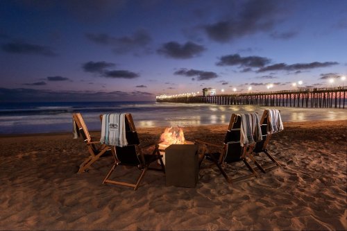 A cozy beach scene at dusk with chairs around a fire pit, overlooking a pier illuminated by lights.
