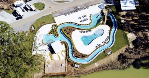 Aerial view of a winding pool area with slides, surrounded by lounge chairs and greenery near a water body.