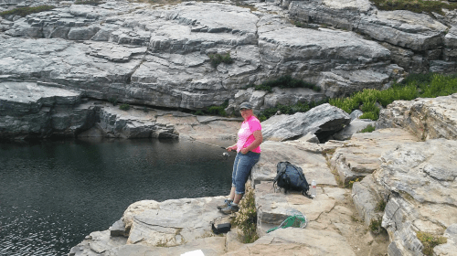 A person in a pink shirt stands on rocky terrain by a calm body of water, fishing with a rod.