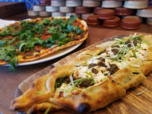 A wooden board with a savory flatbread topped with herbs and cheese, alongside a pizza garnished with fresh greens.