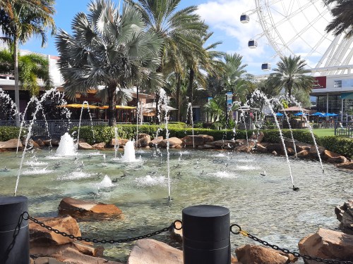 A scenic view of a fountain with water jets, surrounded by palm trees and rocks, near a large Ferris wheel.