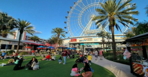 A lively park scene with people relaxing on grass, palm trees, and a large Ferris wheel in the background.