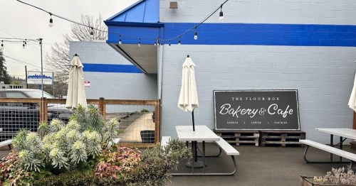 Exterior of The Flour Box Bakery & Cafe, featuring outdoor seating, plants, and a blue building with a sign.
