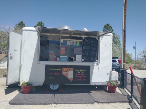 A food truck with a menu displayed, parked in a sunny area with potted plants and a car in the background.