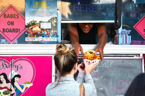 A woman receives a burger and fries from a food truck window, with colorful signs in the background.