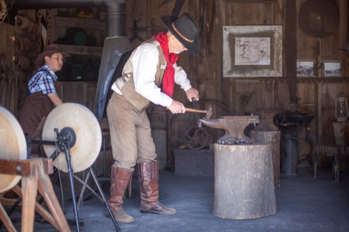 A blacksmith works at an anvil in a rustic workshop, while a boy observes nearby. Tools and equipment are visible around them.