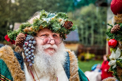 A cheerful man with a long white beard, wearing a festive crown of greenery and ornaments, smiles warmly at the camera.