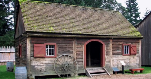 A rustic wooden cabin with a mossy roof, red shutters, and a wagon wheel, surrounded by trees and a wooden fence.