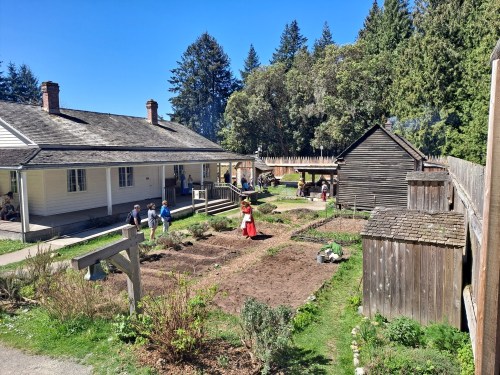 A historic garden scene with people tending to plants, surrounded by buildings and trees under a clear blue sky.