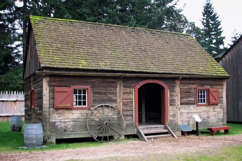 A rustic wooden cabin with a mossy roof, red shutters, and a wagon wheel, surrounded by trees and grass.