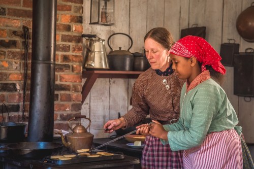 A woman and a girl cook pancakes together on a stove in a rustic kitchen, surrounded by vintage cookware.