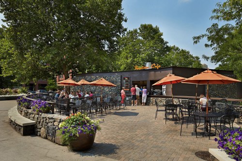 Outdoor café with stone walls, orange umbrellas, and patrons enjoying a sunny day surrounded by greenery and flowers.