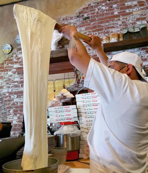 A chef stretches a large piece of dough in a pizzeria, with pizza boxes stacked in the background.