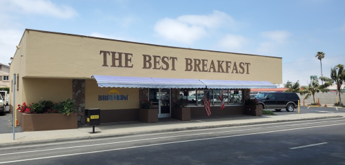 A restaurant building with a sign reading "The Best Breakfast," featuring outdoor seating and a sunny sky.