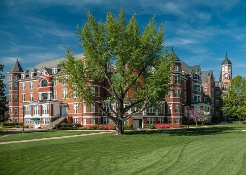 Historic brick building with a large tree in front, set against a blue sky and green lawn.
