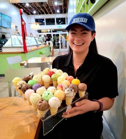 A smiling woman in a cap holds a tray of colorful ice cream cones in a vibrant ice cream shop.
