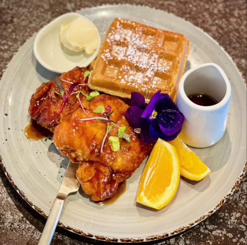 A plate featuring fried chicken with syrup, a waffle, orange slices, a small jug of syrup, and a flower garnish.