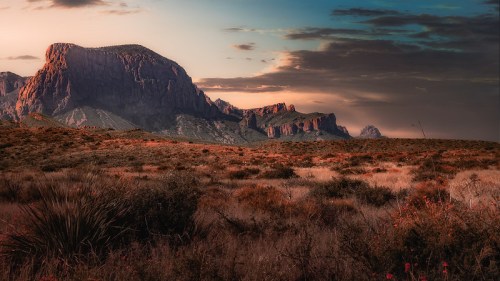 A scenic view of rugged mountains under a colorful sky, with a foreground of dry grass and sparse vegetation.