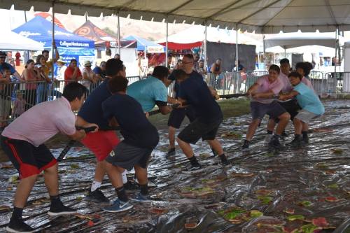 A group of young men participate in a tug-of-war on a wet, slippery surface, surrounded by tents and spectators.