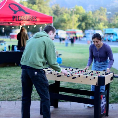 Two people play foosball outdoors at an event, with food trucks and a red tent in the background.