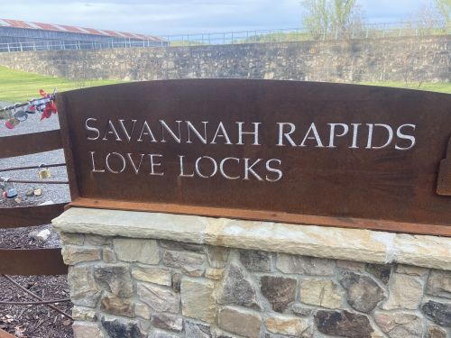 Sign reading "Savannah Rapids Love Locks" on a stone and metal structure, with a scenic background.