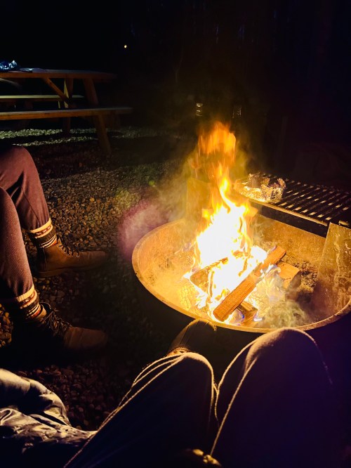 A cozy campfire burns brightly at night, with two pairs of legs visible nearby, surrounded by gravel and picnic tables.