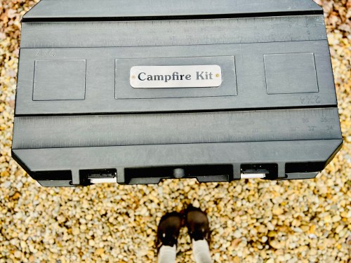 A person holding a black campfire kit above their feet, standing on a gravel surface.