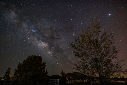 A starry night sky featuring the Milky Way, with trees and mountains silhouetted in the foreground.