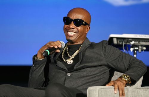 A smiling man in sunglasses and a black suit sits on a stage, holding a microphone and wearing gold jewelry.