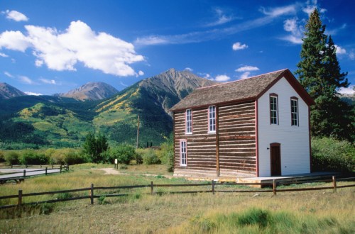 A rustic wooden house stands in a grassy field, surrounded by mountains and a blue sky with fluffy clouds.