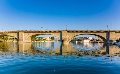 A stone bridge spans a calm river, reflecting the clear blue sky and surrounding greenery.
