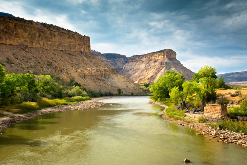 A serene river flows between rocky cliffs, surrounded by lush greenery under a cloudy sky.