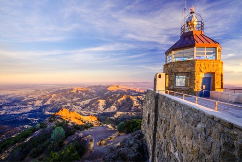 A scenic view of a stone lighthouse on a hill, overlooking rolling hills and valleys at sunset.